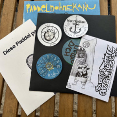 Eine schwarze LP-Hülle mit dem Etikett der Seite A liegt auf dem Cover von Paddelnohnekanu „niemand liebt dich (mehr)“. Auf der schwarzen Innenhülle liegen drei Aufkleber von paddelnohnekanu und dem Label Krachige Platten und ein Zine von der Rattster Review Story. Darauf ist ein teilm der Ilklustration zu sheen mit einem Hamster, der sich die Augen zuhält und dem Schriftzug „Rattster“.