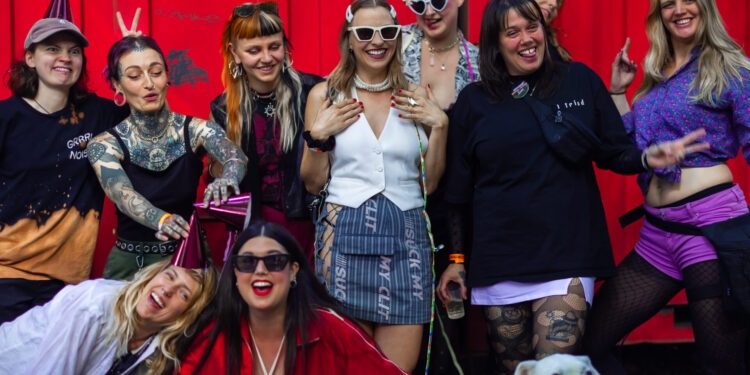 Vor einem roten Hintergrund sieht man die Mitglieder des Grrrl Noisy Kollektivs stehen, ein Hund ist mit auf dem Bild