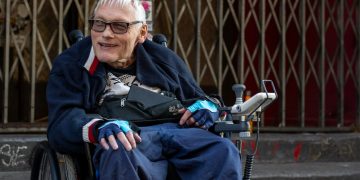 Das Bild zeigt einen Rollstuhlfahrer vor einer Gitterpforte mit Stufen. An der Pforte hängt ein handgeschriebenes Bild, darauf steht, dass ein Konzert von The Selecter ausfällt.