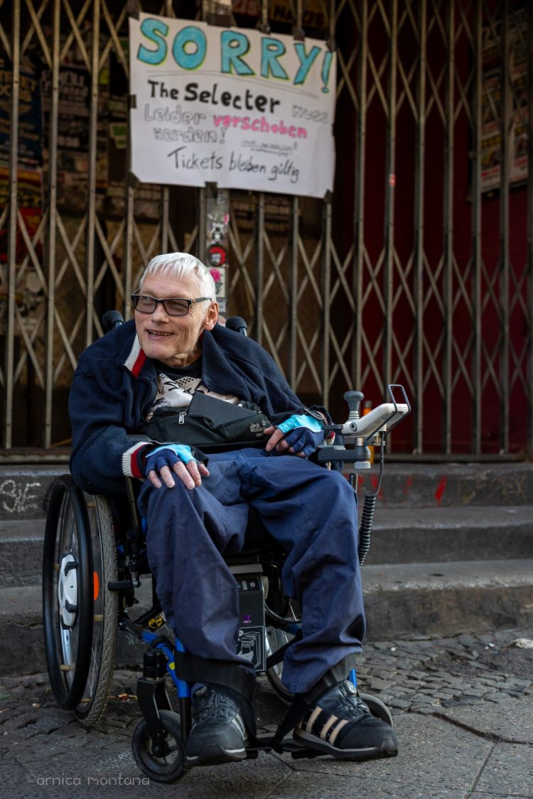 Das Bild zeigt einen Rollstuhlfahrer vor einer Gitterpforte mit Stufen. An der Pforte hängt ein handgeschriebenes Bild, darauf steht, dass ein Konzert von The Selecter ausfällt.