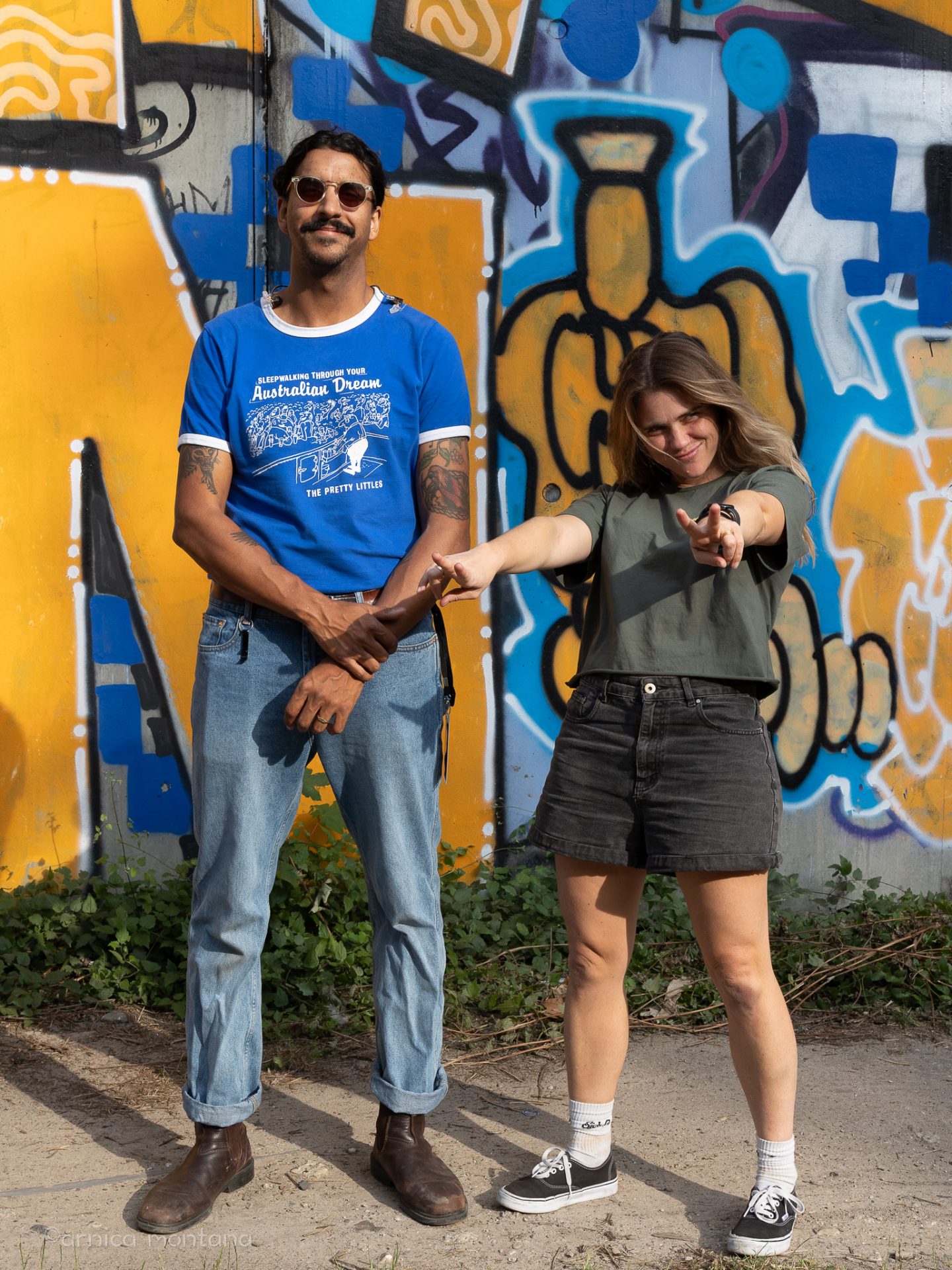Vor einer gelb-blauen Graffitiwand stehen Natalie und Greg von Press Club. Natalie streckt lässig zurückgelehnt die Hände nach vorne und Greg trägt eine Sonnenbrille.