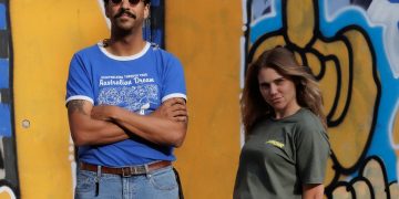 Vor einer gelb-blauen Graffitiwand stehen Natalie und Greg von Press Club. Natalie hält lässig zurückgelehnt die Hände hinterm Rücken und Greg trägt eine Sonnenbrille.