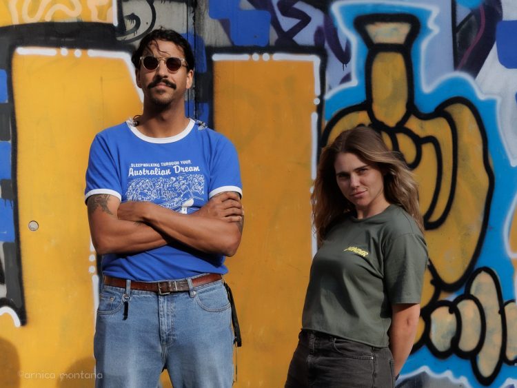 Press Club & DZ Deathrays 1 Vor einer gelb-blauen Graffitiwand stehen Natalie und Greg von Press Club. Natalie hält lässig zurückgelehnt die Hände hinterm Rücken und Greg trägt eine Sonnenbrille.