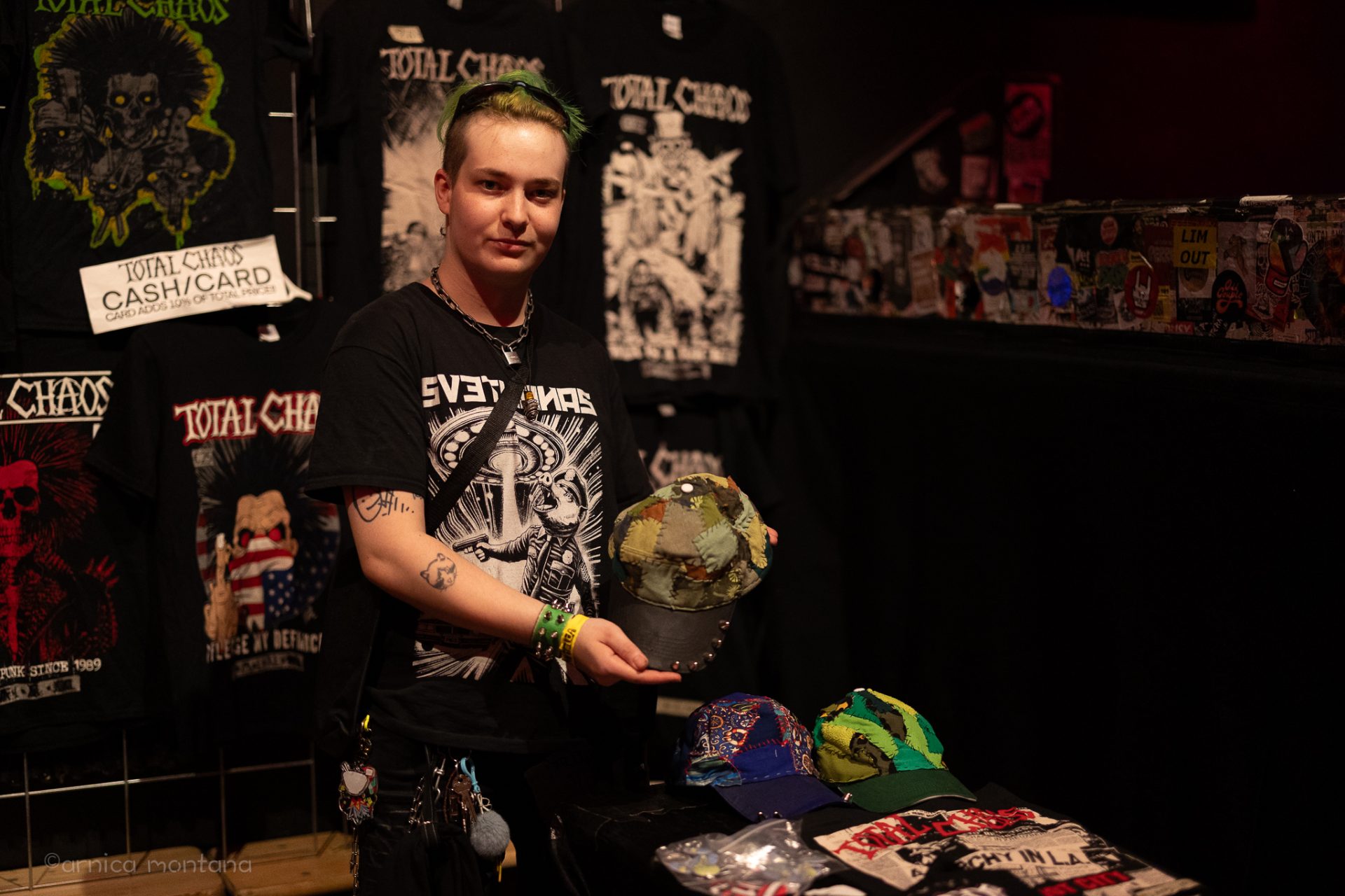 Ein Künstler mit grünen Haaren hält eine DIY-Mütze ins Bild. Im Hintergrund sind T-shirts und Plakate von Total Chaos zu erkennen. Er trägt ein T-Shirt mit der Aufschrift "Svetlanas".