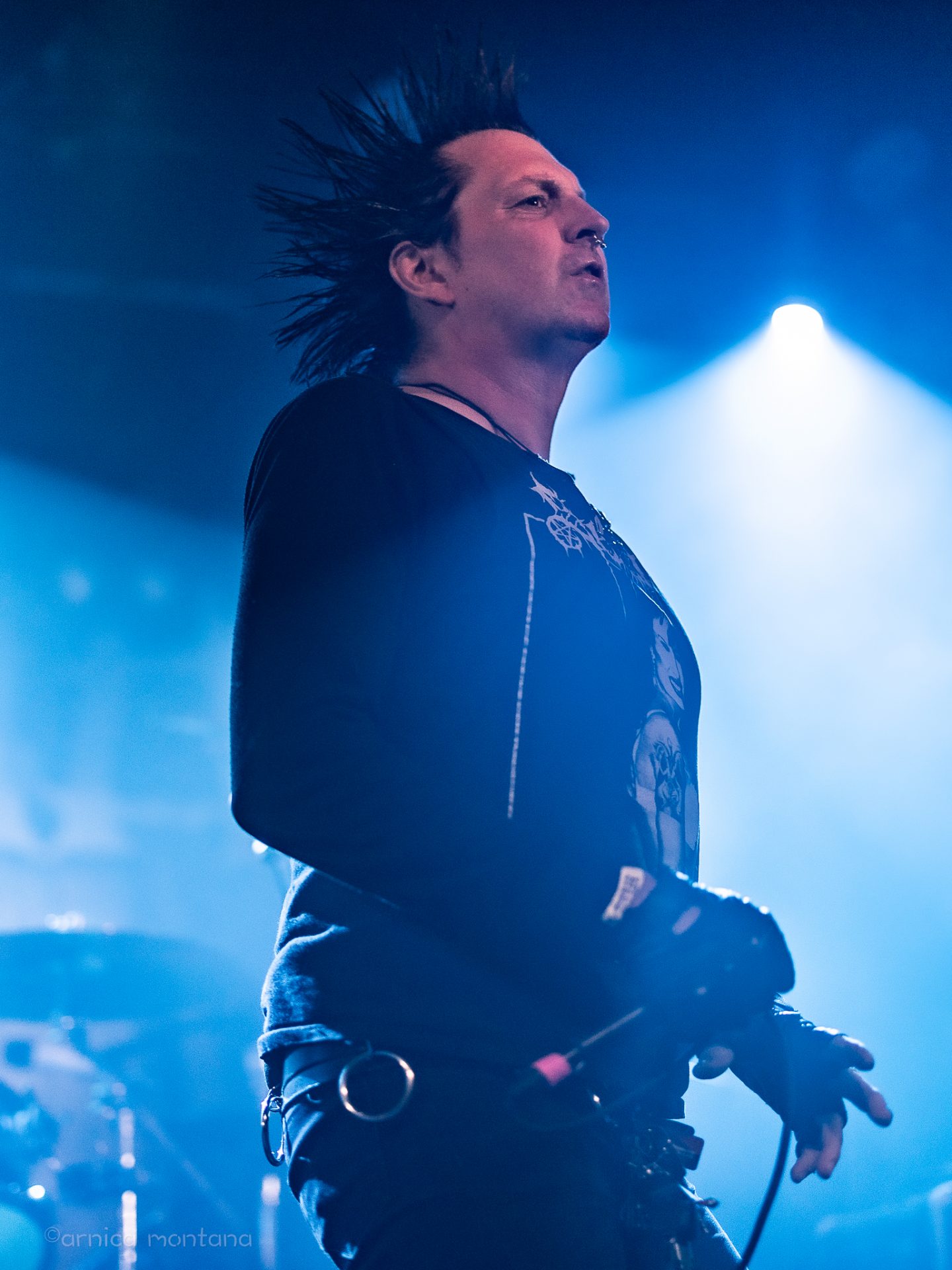 Diversity Dive #14 meets MusInclusion #33 - Total Chaos & Rumkicks & Agrotóxico 12 Ein Mann mit schwarzer Spike-Frisur hält eine Mikrofon in der Hand. Er trägt schwarze Kleidung. Das Bühenlicht im Hintergrund ist bläulich und lässt Teile eines Schlagzeugs erahnen..