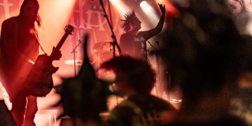 Eine lebendige Konzertszene mit rötlichen Lichtern. auf der Bühne sind mehrere Musiker zu erkennen, im Vordergrund die Personen sind unscharf und haben Spike-Frisuren.