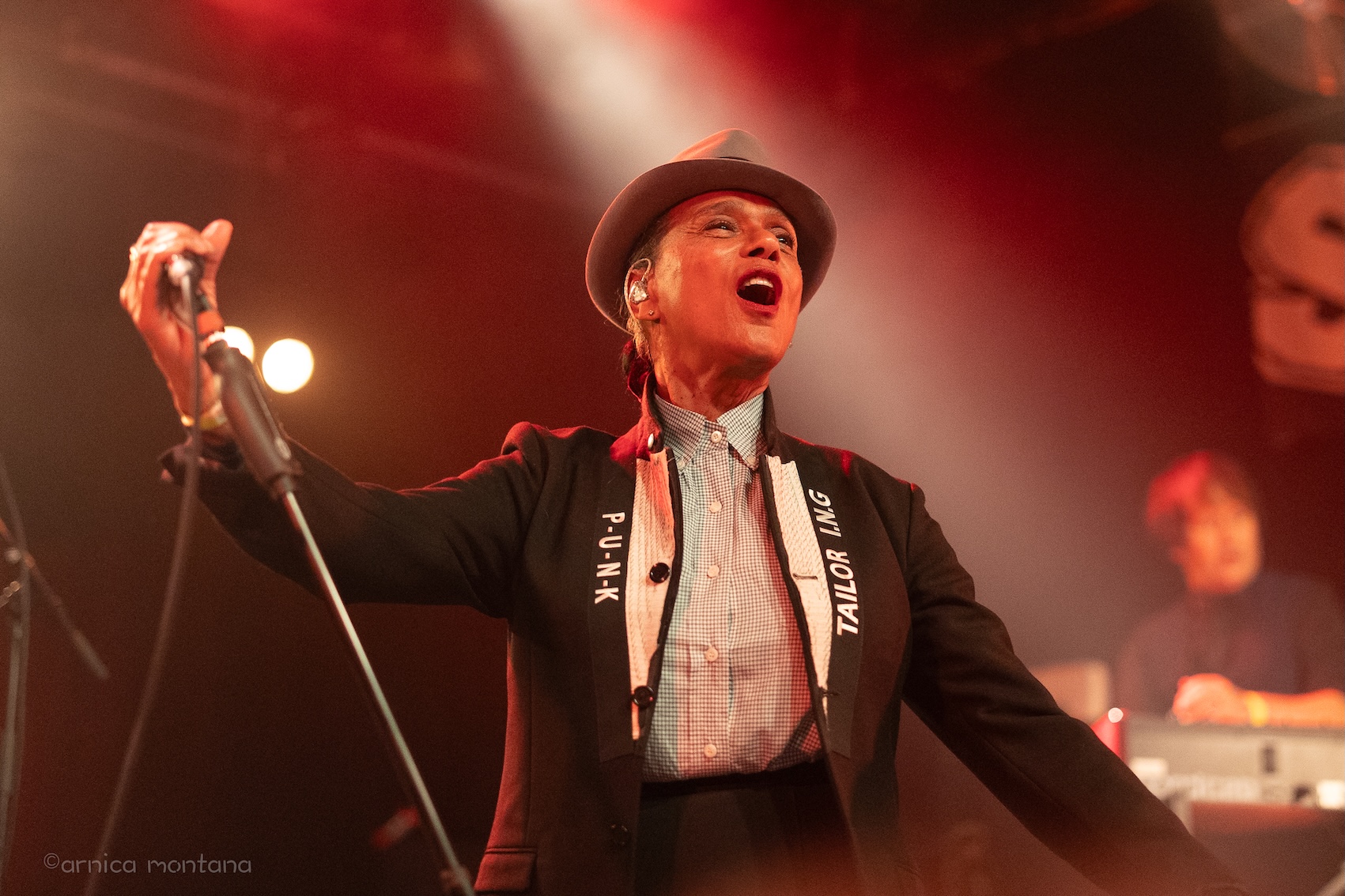 Pauline Black hält das Mikrofon schräg zur Seite gekippt und singt mit einer ausdrucksvollen Pose und geöffnetem Mund. Das Bühnelicht ist rot mit einem weißen Schlaglicht, das auf die Sängerin gerichtet ist.
