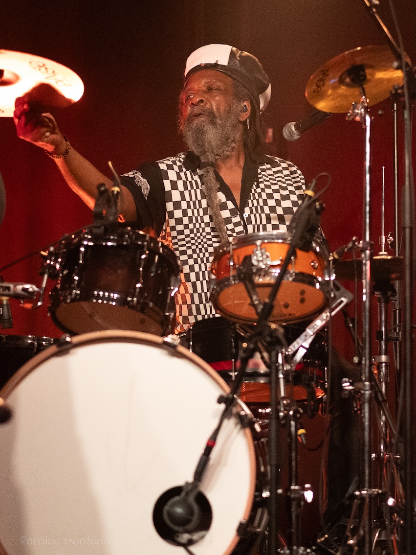 Ein Drummer mit Bart und Dreadlocks dämpft mit der rechten Hand ein Becken am Schlagzeug, in der linken Hand hält er die Drumsticks. 