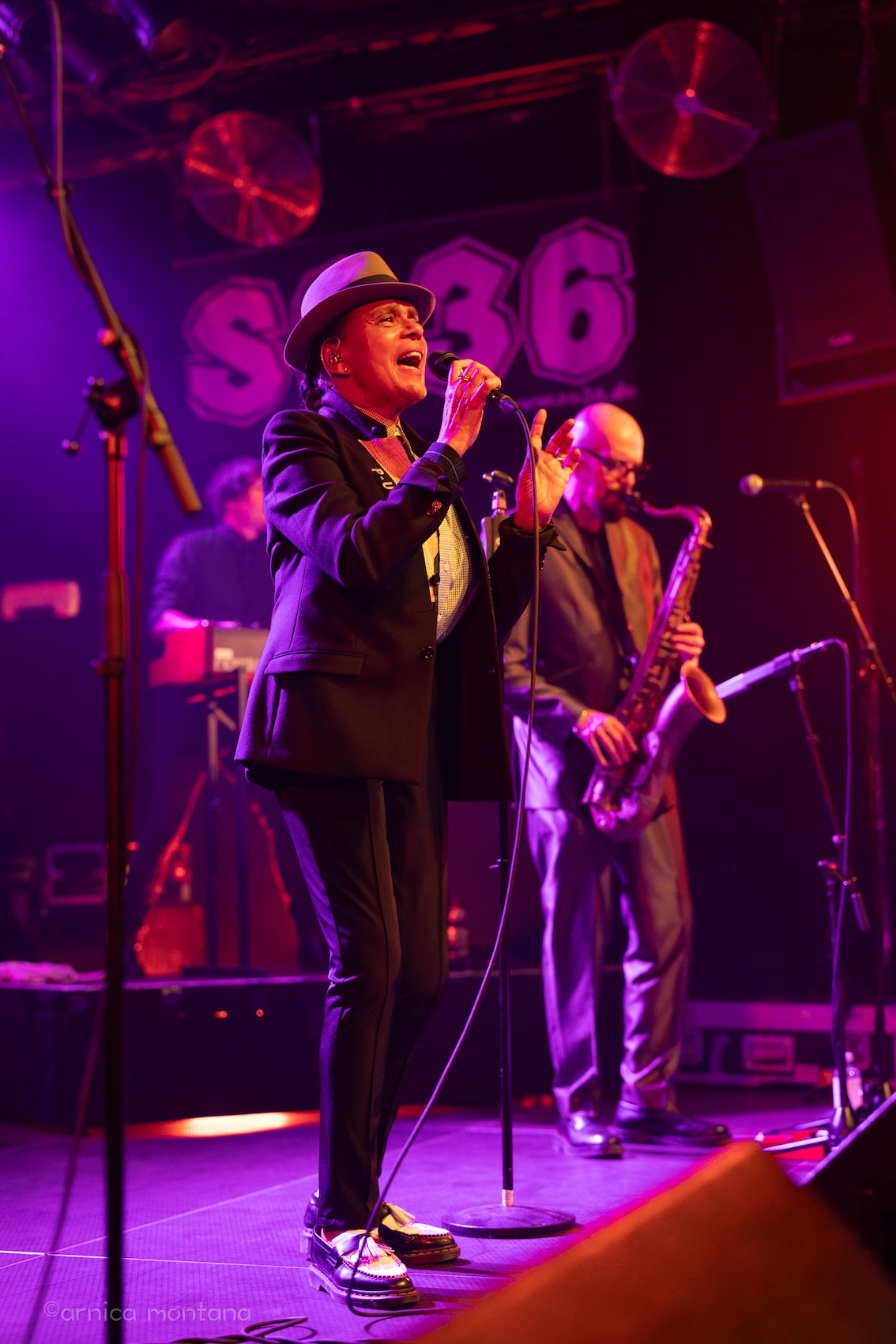 Pauline Black in typischem Bekleidungsstil singt in das Mikrophon, rechts daneben der Saxophonist, im Hintergrund der Keyboarder. Auf der in Pink-Violettes Licht getauchten Bühne sind die Buchstaben SO36 zu erkennen.