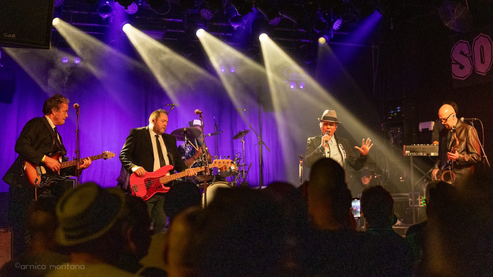 Eine Bühne ausgeleuchtet in violettem Licht, zeigt in Schlaglichtern die Band zusammen mit Pauline Black. Links stehen der Gitarrist und der Bassist, rechts der Saxophonist und ein Keyboard, im Hintergrund ist der Schlagzeuger zu erkennen.