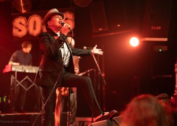 Diversity Dive #15 - The Selecter - Pauline Black: A 2-Tone Story 2 Eine Frau mit Pork Pie Hut in schwarz mit weißem Hutband, schwarzem Jacket und schwarzen Cigarette Pants steht mit einem Schuh auf dem Monitor und singt zum Publikum. Eine Hand macht eine einladenden Bewegung, die andere Hand hält das. Mikrofon, während sie singt.