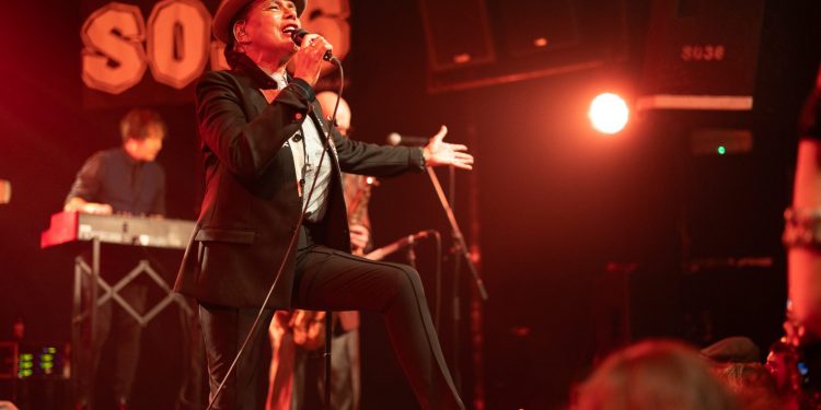 Diversity Dive #15 - The Selecter - Pauline Black: A 2-Tone Story 1 Eine Frau mit Pork Pie Hut in schwarz mit weißem Hutband, schwarzem Jacket und schwarzen Cigarette Pants steht mit einem Schuh auf dem Monitor und singt zum Publikum. Eine Hand macht eine einladenden Bewegung, die andere Hand hält das. Mikrofon, während sie singt.