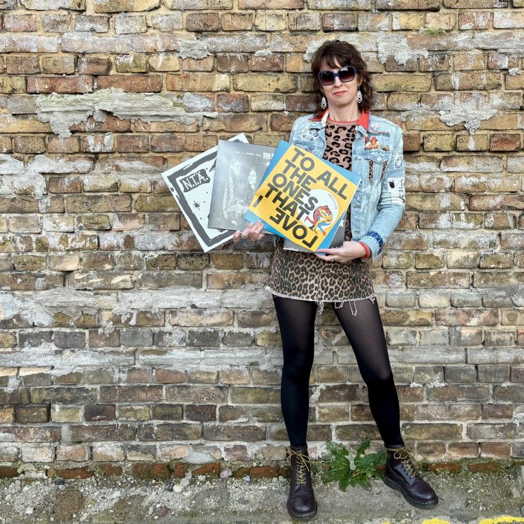 Eine Frau mit mit Jeans Jacken und selbstgestickten Aufnähern hält drei Plattenalben. Der Hintergrund ist eine Backsteinmauer.