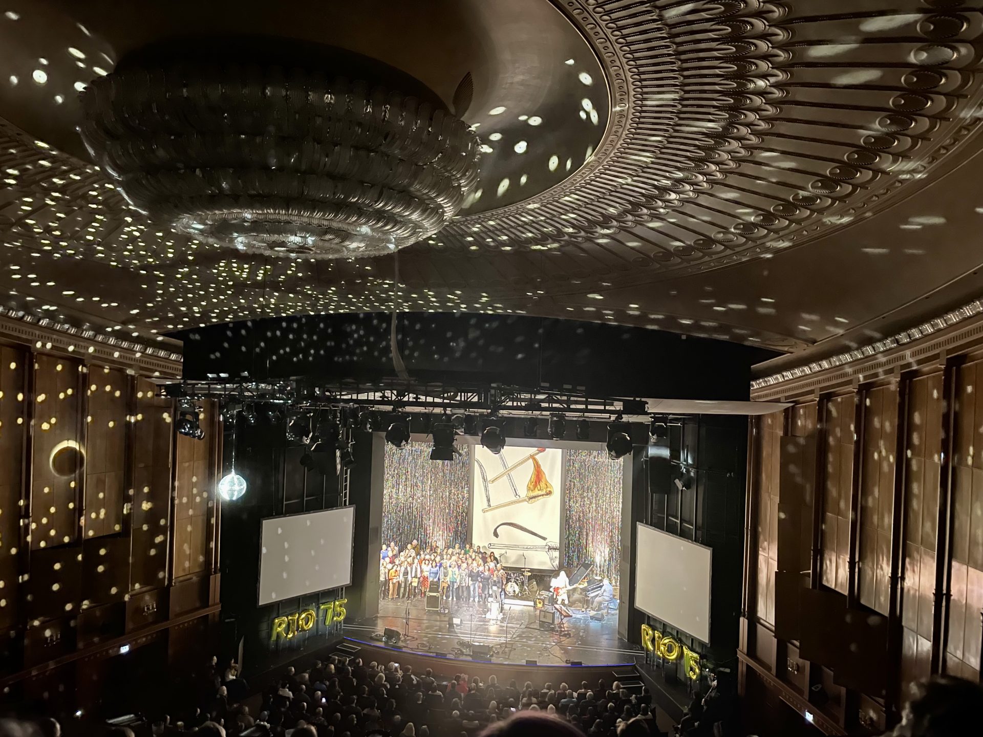 Blick auf die Bühne mit einem Glittervorhang, der rot angestrahlt ist und einem großen Banner als Bühnenbild, das einige gezeichnete Streichhölzer zeigt, die bereits abgebrannt, unverbraucht oder frisch entzündet sind. Links neben dem Bühnenbild stehen viele verschiedene Personen und Instrumente. Davor steht eine Frau mit einer Gitarre und rechts daneben sitzt ein mann am Flügel. Der Saal wird durch eine angestrahlte Discokugel links in helle Lichtreflexe getaucht. Das Publikum auf Sitzplätzen ist undeutlich von hinten zu sehen. links und rechts vor der Bühne sind zwei Leinwände unter denene in goldenen, aufblasbaren Buchstaben RIO 75 steht.