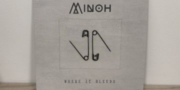 Ein Plattencover mit zwei geometrisch angeordneten Sicherheitsnadeln und einem ebenfalls geometrisch stilisierten Bandnamen "Minoh" ist zu sehen auf einer hölzernen Unterlage vor einer weißen Wand. Das Albumcover ist zweifarbig in grau mit schwarzer Schrift gehalten und wirkt aufgeräumt.
