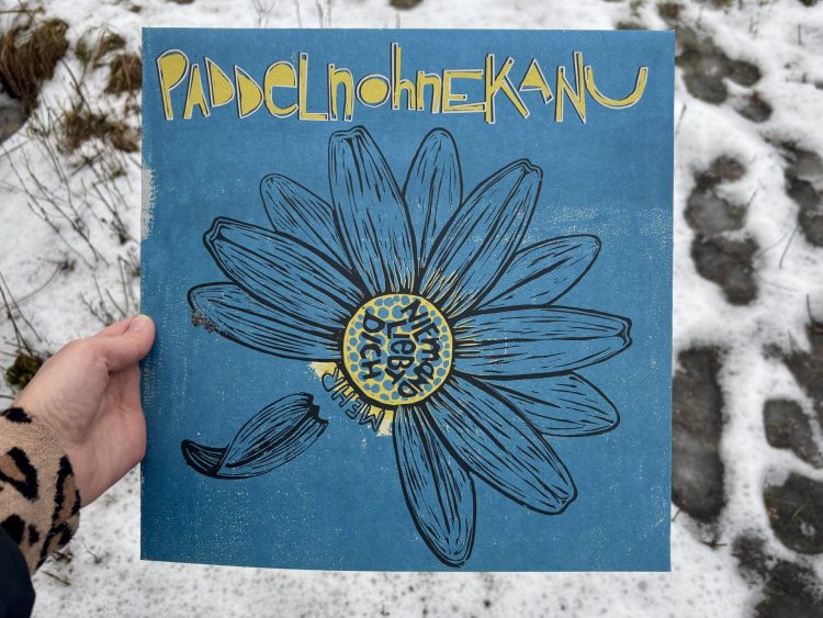 paddelnohnekanu - Niemand liebt dich (mehr) / Rattster Staffel 3, Folge #1 1 Ein Albumcover mit eines Linolschnitts eines Gänseblümchens auf blauem Grund. Ein paar Blütenblätter fehlen, eines löst sich gerade vom Blütenstandsboden. Die inneren, gelben Röhrenblüten sind angedeutet in gelb auf den blauen Untergrund gedruckt. Der Schriftzug paddelnohnekanu ist gelb mit weißer Umrandung prominent am oberen Rand platziert. Daraus ergibt sich eine Farbpalette von blau, gelb, weiß und schwarz für das Cover. Eine Hand hält das Album vor einem Hintergrund aus Schnee mit Fußspuren.