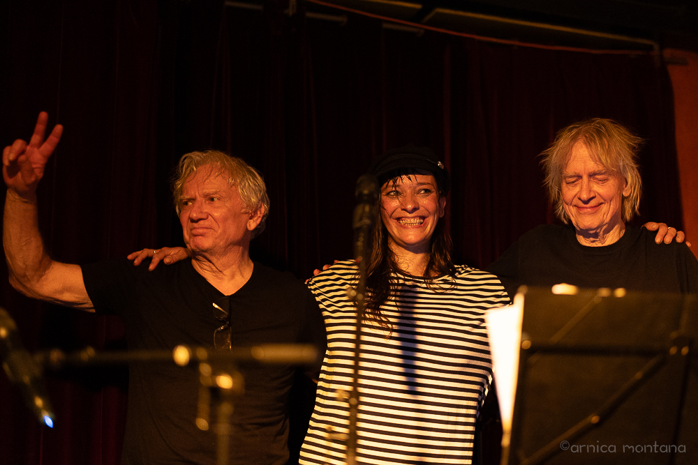 Drei Personen stehen Arm in Arm auf einer Bühne. Sie sehen entsapnnt und glücklich aus. Im Vordergrund sind ein Notenständer und ein Mikrofonständer zu erkennnen, der Hintergrund ist ein schwarzer Bühnenvorhang.