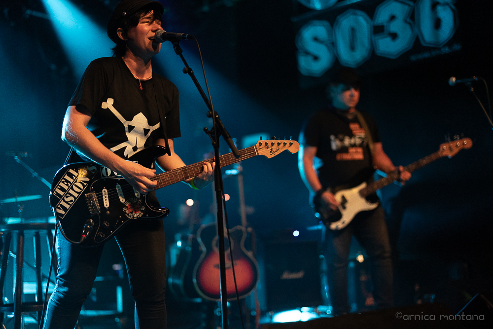 Eine Bühne mit dem Schriftzug SO36 m Hintergrund ist zu sehen. Das Bühnenlicht ist ein petrolblau, die beiden Personen mit Instrumenten sind in natürliches Licht getaucht und heben sich farblich aus dem blauen Licht ab. Im Vordergrund llinks spielt eine Frau mit einem schwarzen T-Shirt mit weißem Print eine Gitarre, links spielt ein Mann im Hintergrund einen E-Bass.