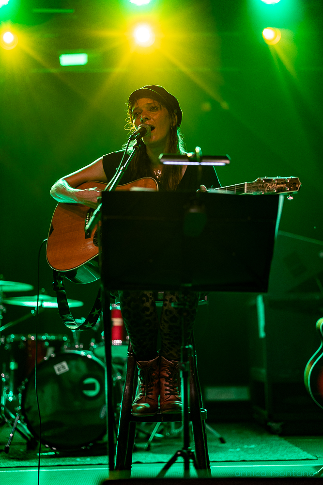 Eine Frau mit einer Akstikgitarre sitzt auf einem Barhocker hinter einem Notenständer auf der Bühne. Sie trägt eine dunkle Schirmmütze und braune Boots. Im Hintergrund ist ein Schlagzeug zu erkennen. Die Szene ist in grün-gelbes Licht getaucht.