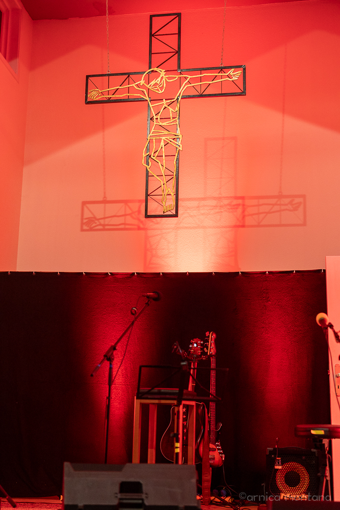 Ein Kreuz aus montierten Metallstreben präsentiert eine Jusus-Figur aus hellen Leuchtröhren. darunter stehen die Musikinstrumnete von Ton Steine Scherben und Birte Volta. Die Szenen ist in euchtendes, rötliches Licht getaucht.