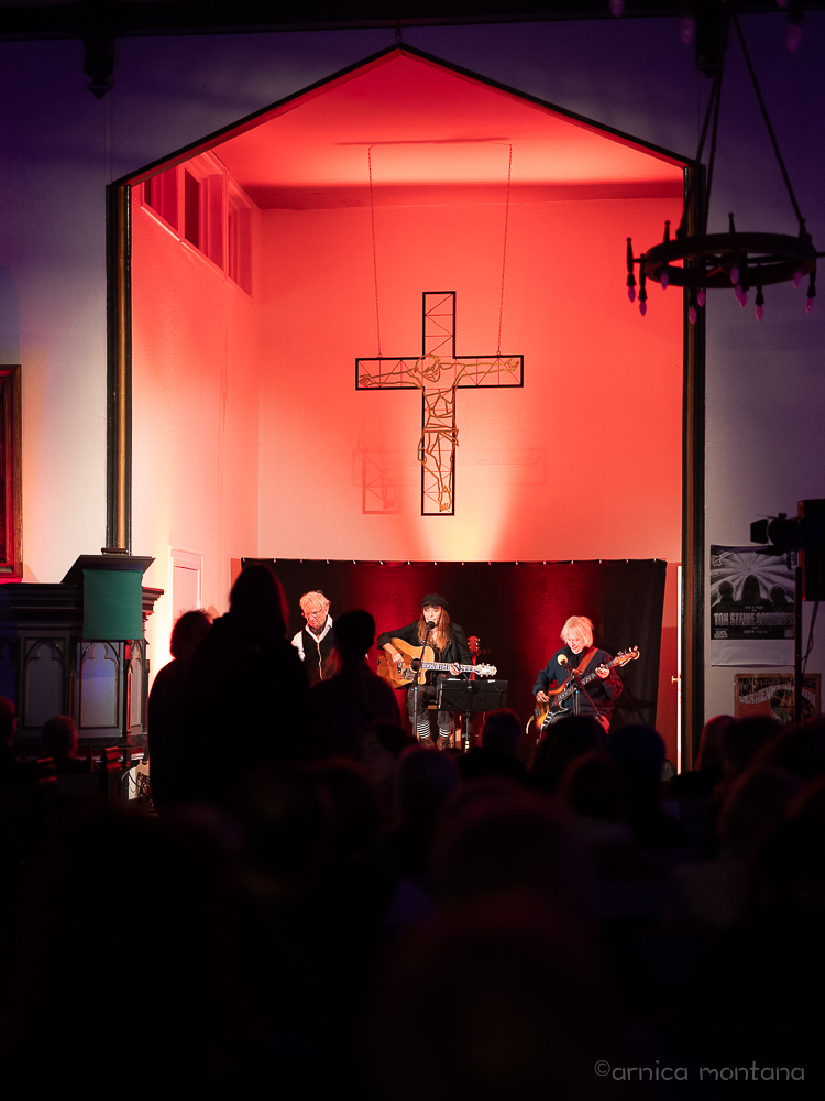 Drei Personen sitzen zusammen unter einem Jesus am Kreuz in einem abgeteilten Raumstück als Bühne. Daneben sind ein links ein Kanzel und rechts oben ein Adventskranz zu erkennen. Unter dem Adventskranz annder <wand hängt ein Ton Steine Scherben Tourplakat. Im Vordergrund sind Silhouetten vom Publikum zu erkennen. Der Raum ist in rötliches bis violette Licht getaucht und vermitelt eine Konzertathmosphäre.. 