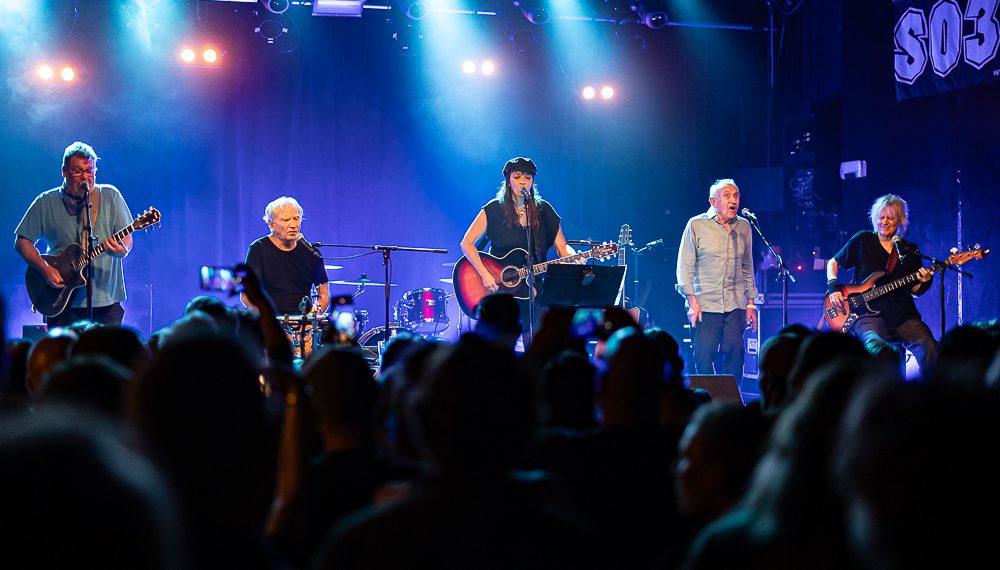 Fünf Personen stehen auf einer blau erleuchtete Bühne mit unterschiedlichen Instrumenten. Im Vordergrund ist das Publikum von hinten zu erkennen.