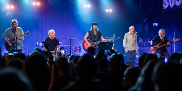 Fünf Personen stehen auf einer blau erleuchtete Bühne mit unterschiedlichen Instrumenten. Im Vordergrund ist das Publikum von hinten zu erkennen.