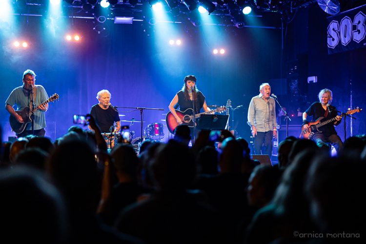 Fünf Personen stehen auf einer blau erleuchtete Bühne mit unterschiedlichen Instrumenten. Im Vordergrund ist das Publikum von hinten zu erkennen.
