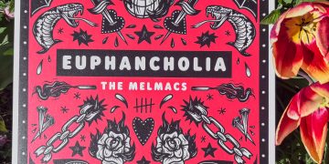 Ein Musikalbum wird neben Blumen und Grünpflanzen präsentiert, Es ist rot und zeigt schwarz-weiße Illustrationen.
