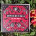 Ein Musikalbum wird neben Blumen und Grünpflanzen präsentiert, Es ist rot und zeigt schwarz-weiße Illustrationen.