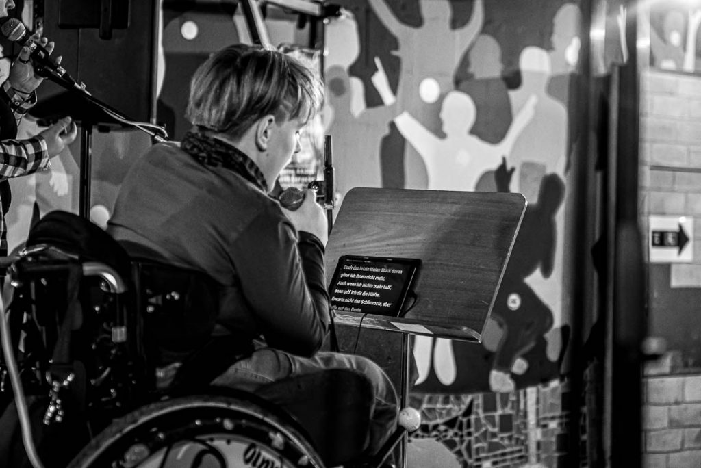 MusInclusion #34 - Band Workshop und so viele weitere coole Dinge! 2 Eine jugendliche Person im Rollstuhl vor einem Notenständer schaut auf einen Bildschirm, der einen Text anzeigt. Im Hintergrund ist ein Mikrofon zu sehnen und eine Wandgestaltung mit Kindersilhouetten.