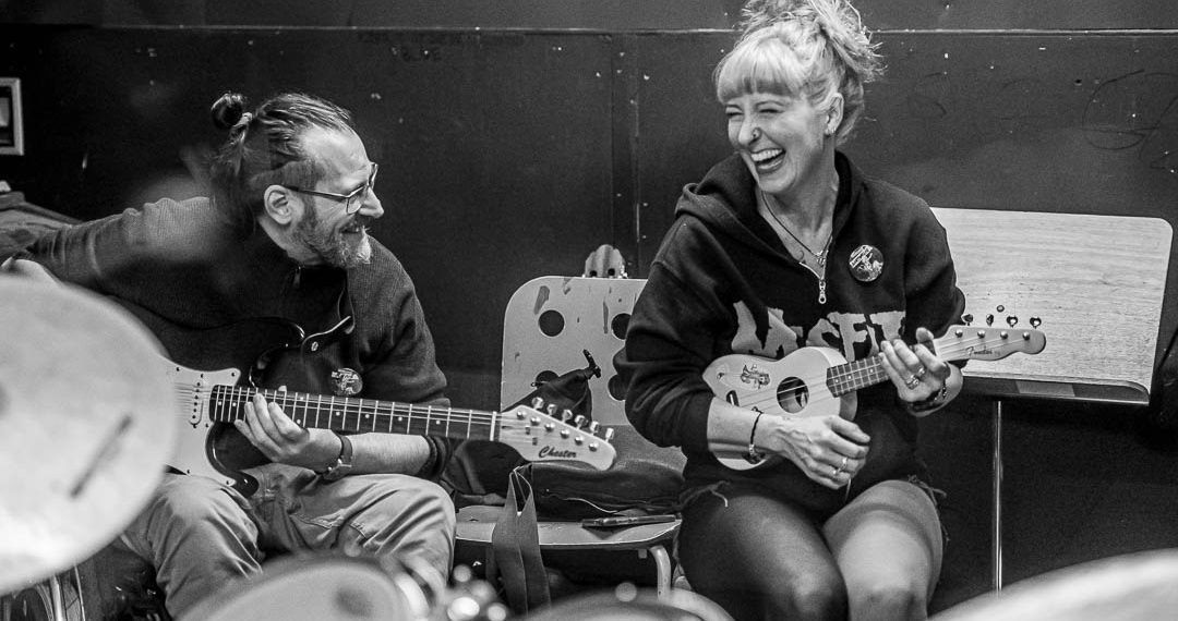 Zwei Personen mit Musikinstrumenten, eine Ukulele und eine Gitarre; sitzen zusammen und lachen.