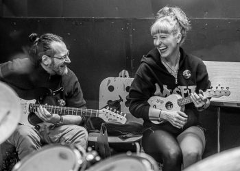 Zwei Personen mit Musikinstrumenten, eine Ukulele und eine Gitarre; sitzen zusammen und lachen.