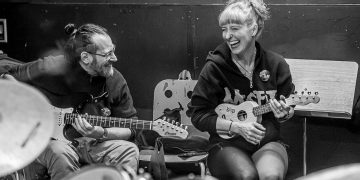 Zwei Personen mit Musikinstrumenten, eine Ukulele und eine Gitarre; sitzen zusammen und lachen.