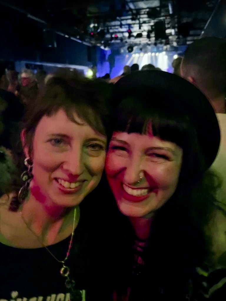 MusInclusion #34 - Band Workshop und so viele weitere coole Dinge! 5 Zwei Personen schauen lachend in die Kamera, im Hintergrund ist eine Konzert-Location zu erkennen.