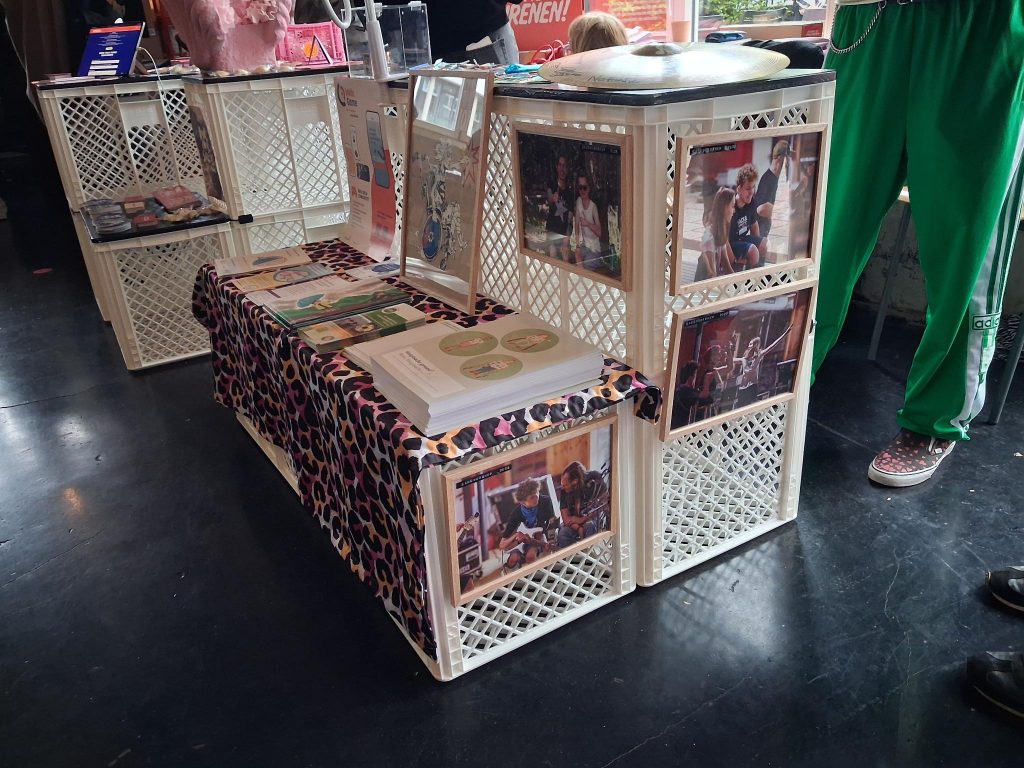 MusInclusion #34 - Band Workshop und so viele weitere coole Dinge! 3 Ein bunter Infostand m it Plakaten, Flyern und Shirts von Musinclusion ist zu sehen.