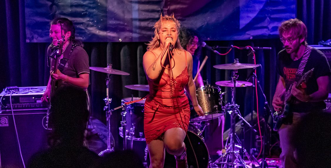 Eine Band steht auf der Bühne, die Sängerin positioniert sich mittig und trägt ein rotes Kleid, im Hintergrund hängt ein Banner mit dem Schriftzug Niranium