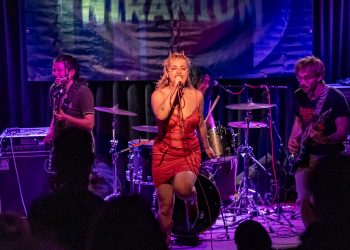 Eine Band steht auf der Bühne, die Sängerin positioniert sich mittig und trägt ein rotes Kleid, im Hintergrund hängt ein Banner mit dem Schriftzug Niranium