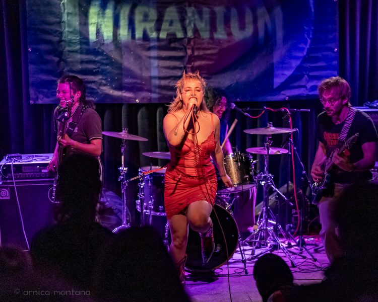 MusInclusion #35 meets Diversity Dive #19 - Niranium Festival 1 Eine Band steht auf der Bühne, die Sängerin positioniert sich mittig und trägt ein rotes Kleid, im Hintergrund hängt ein Banner mit dem Schriftzug Niranium