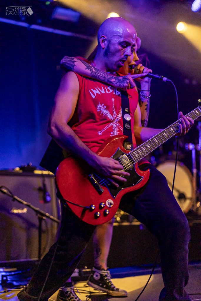 Interview mit Gero / Female Fronted is not a Genre Festival 2026 14 Ein Gitarrist mit einer roten Gitarre und einem roten Bandshirt spielt Gitarre, die Sängerin hat einen Arm um seinen Hals gelegt, macht eine Pose mit dem Zeigefinger und singt in ihr Mikro.