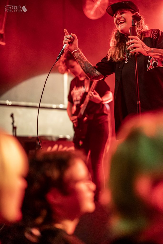 Interview mit Gero / Female Fronted is not a Genre Festival 2026 10 Eine in rötliches Licht getauchte Bühne zeigt zwei Musikerinnen. Eine spielt Girtarre, die andere hält ein Mikrophon, macht eine Zeigefingerpose und lacht.