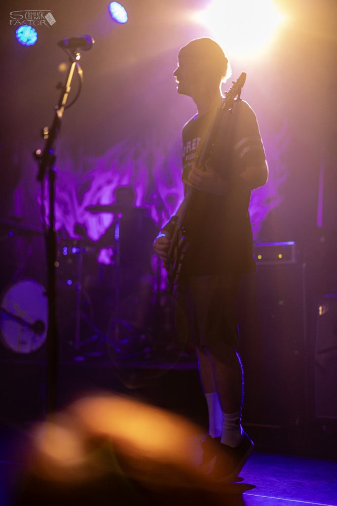 Interview mit Gero / Female Fronted is not a Genre Festival 2026 9 Violettes Licht, ein Goldgelber Lichtschein formt im Gegenlicht die Silhouette des Gitarristen. im Hintergrund ist das Schlagzeug zu erkennen.