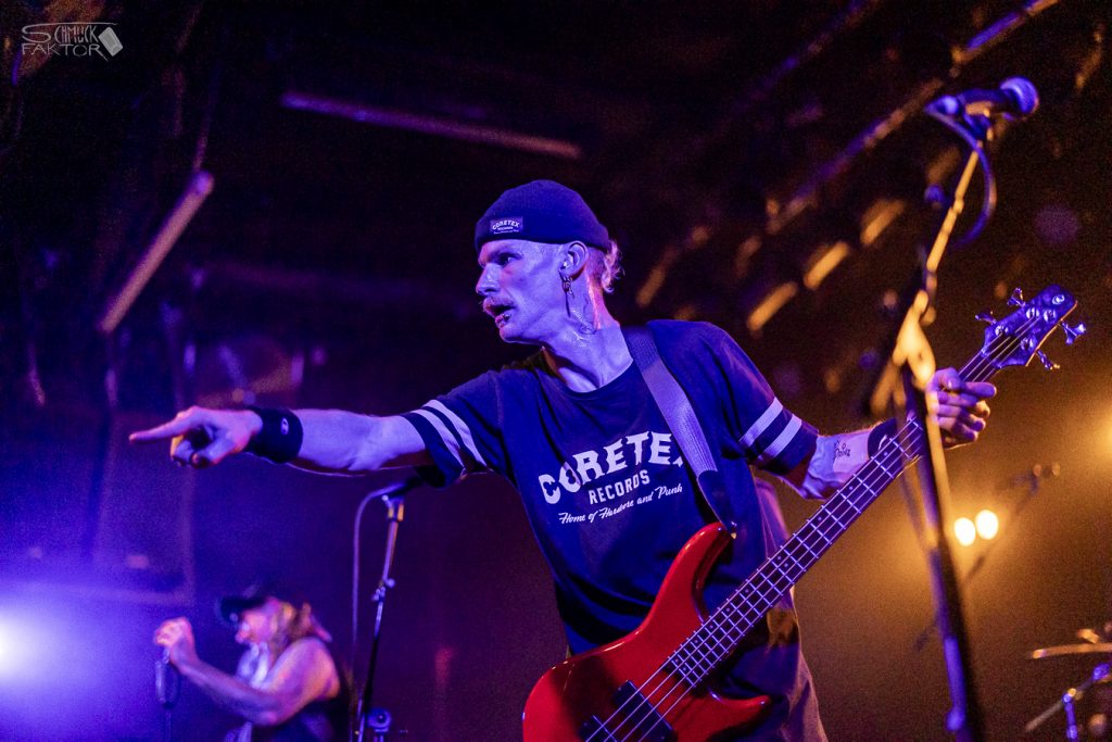 Interview mit Gero / Female Fronted is not a Genre Festival 2026 7 Der Gitarrist macht eine zeigende Pose mit dem Zeigefinger, im Hintergrund ist eine Sängerin zu sehen. Das Bühnenlicht ist violett und goldgelb.