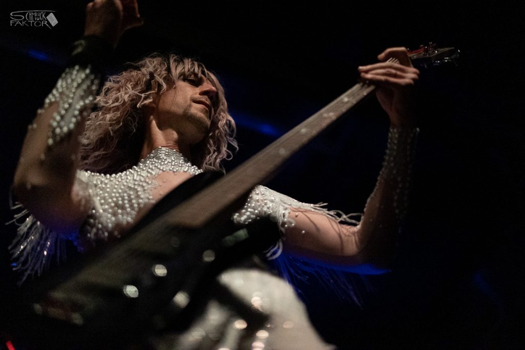Interview mit Gero / Female Fronted is not a Genre Festival 2026 29 Eine Person mit einem hellen, engen Anzug mit Fransen und Perlen besetzt, steht vor dunklem Hintergrund eine Gitarre