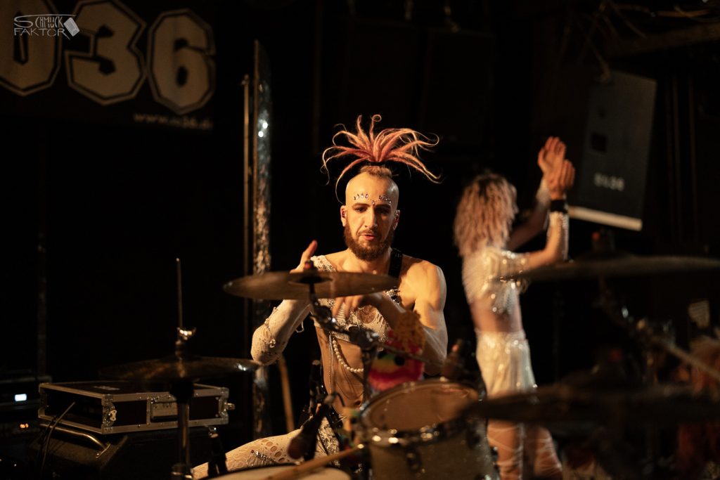 Interview mit Gero / Female Fronted is not a Genre Festival 2026 27 Der Schlagzeuger sitzt vor dunklem Hintergrund, im Hintergrund klatscht eine Person in einem weißen Anzug in beide Hände
