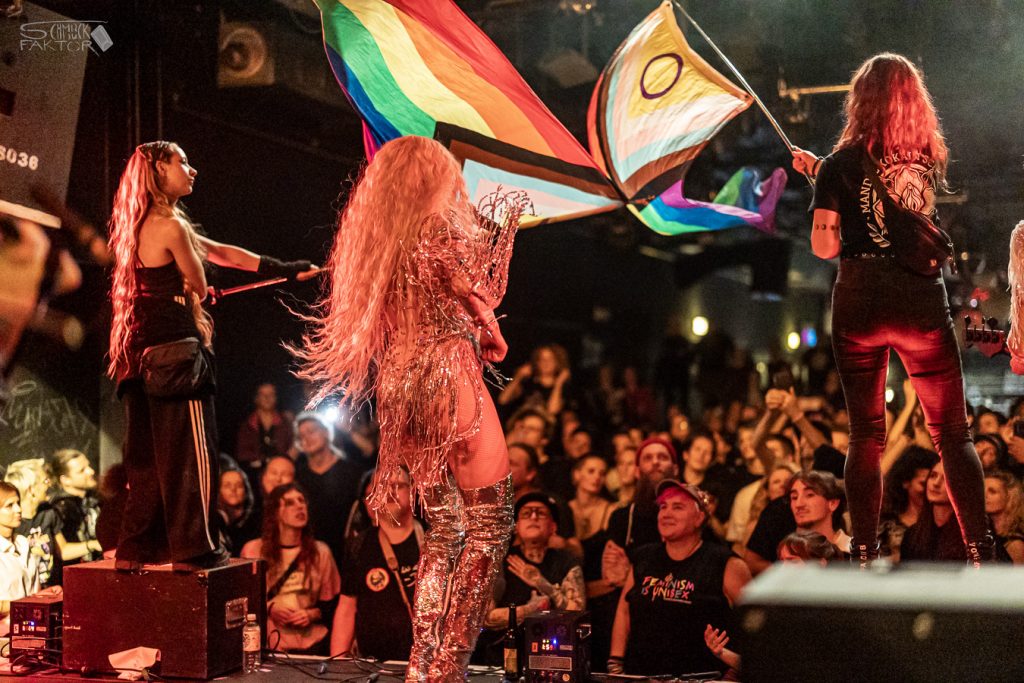 Interview mit Gero / Female Fronted is not a Genre Festival 2026 22 Zwei Personen schwingen bunte Pride Flaggen vor dem Publikum, die Sängerin ist von hinten zu sehen und steht frontal vor dem Publikum